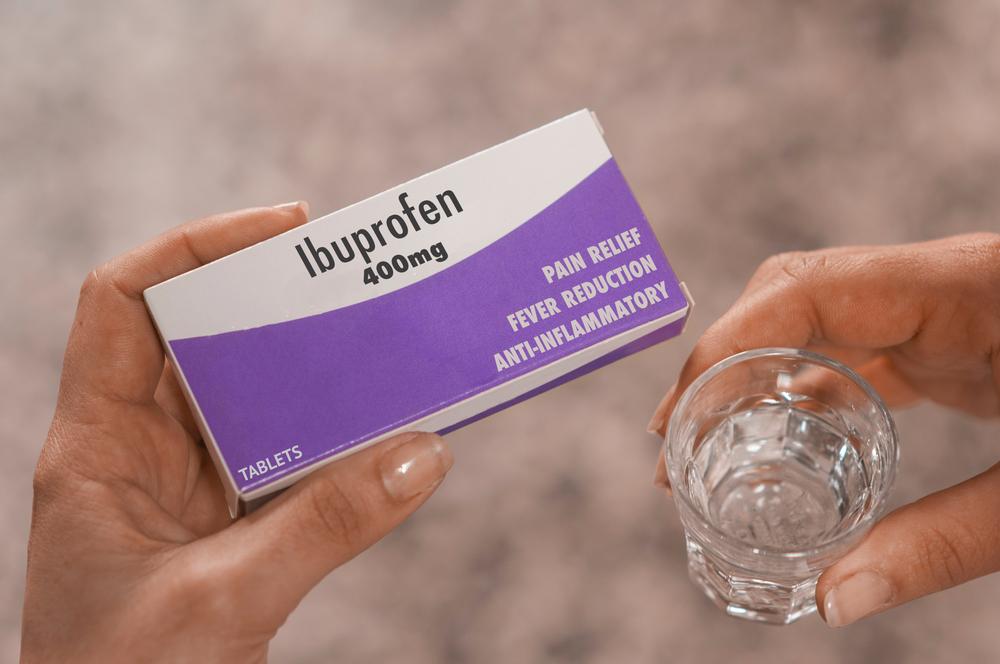 A woman holding a glass of water and a box of Ibuprofen tablets.