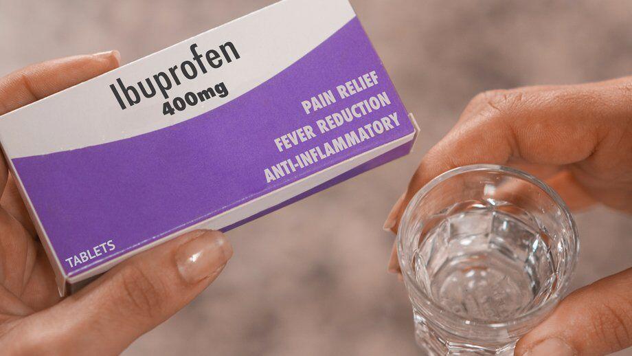 A hand holding a glass of water next to a box of ibuprofen pills.