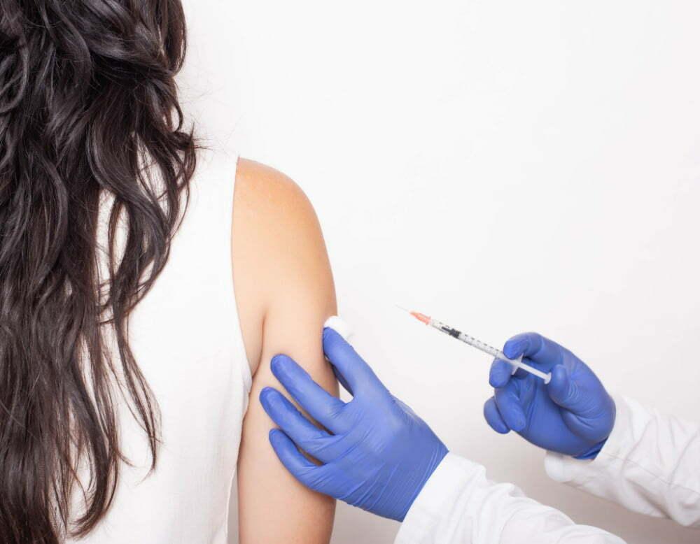 A person in a white lab coat and blue gloves is giving a woman a vaccination in her upper arm.