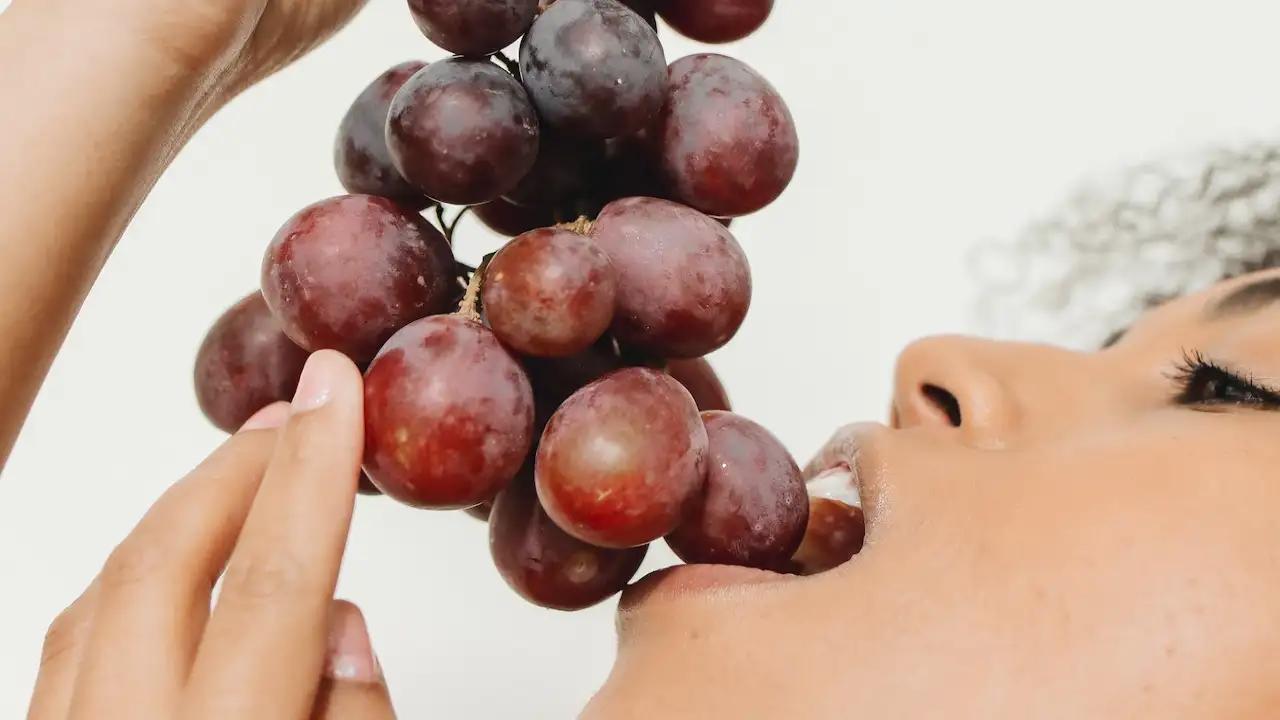 A young woman is eating red grapes, holding a bunch of them in her hand and biting into one.