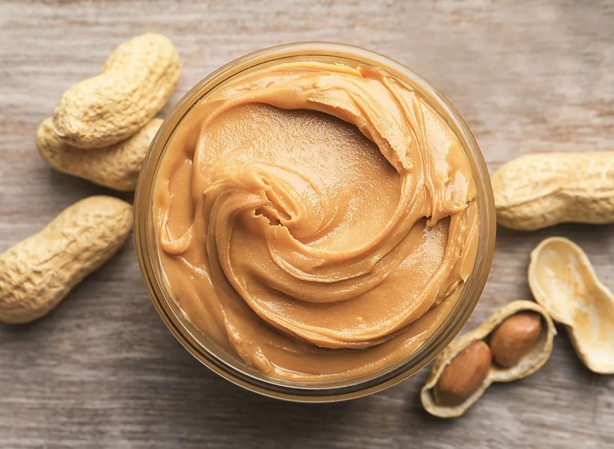 An open jar of peanut butter next to scattered peanuts.