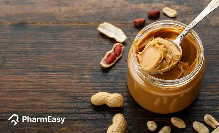 An open jar of peanut butter with a silver spoon in it, next to scattered peanuts in their shells.