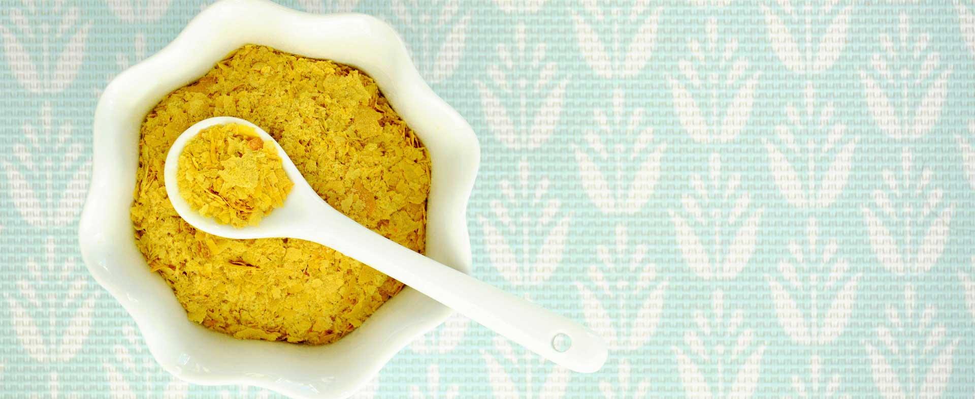 A white bowl filled with nutritional yeast flakes and a white spoon.