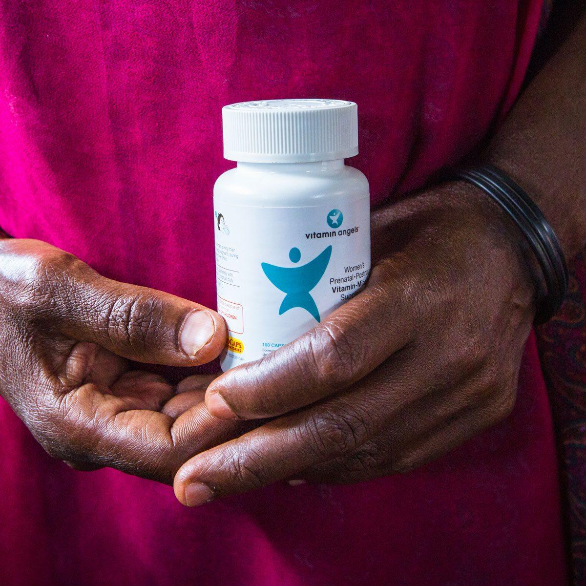 A dark-skinned person is holding a bottle of Vitamin Angels prenatal vitamins in their hands.