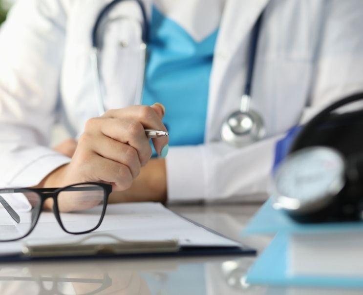 A doctor in a white coat is holding a pen and looking at a clipboard.