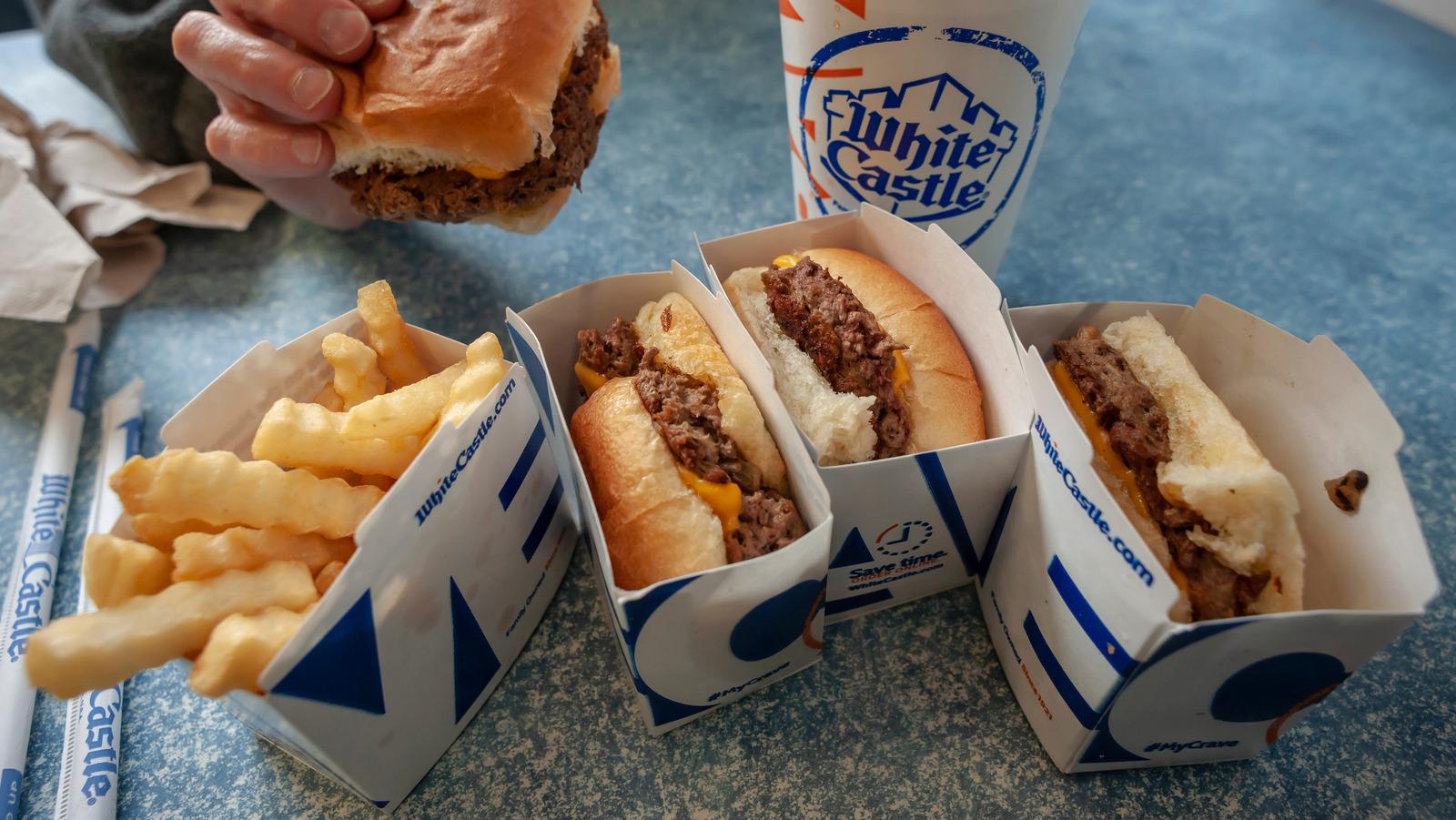 A hand holding a White Castle slider hamburger with a box of three more sliders and an order of fries next to a fountain drink cup.