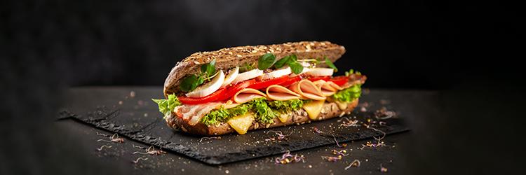 A sandwich with ham, cheese, tomato, lettuce, and sprouts on a whole wheat baguette.