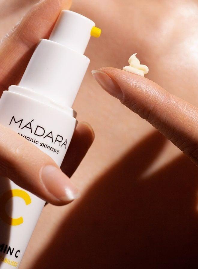 A womans finger is applying a yellow-white cream from a white bottle with an orange cap that has MADARA organic skincare printed on it.
