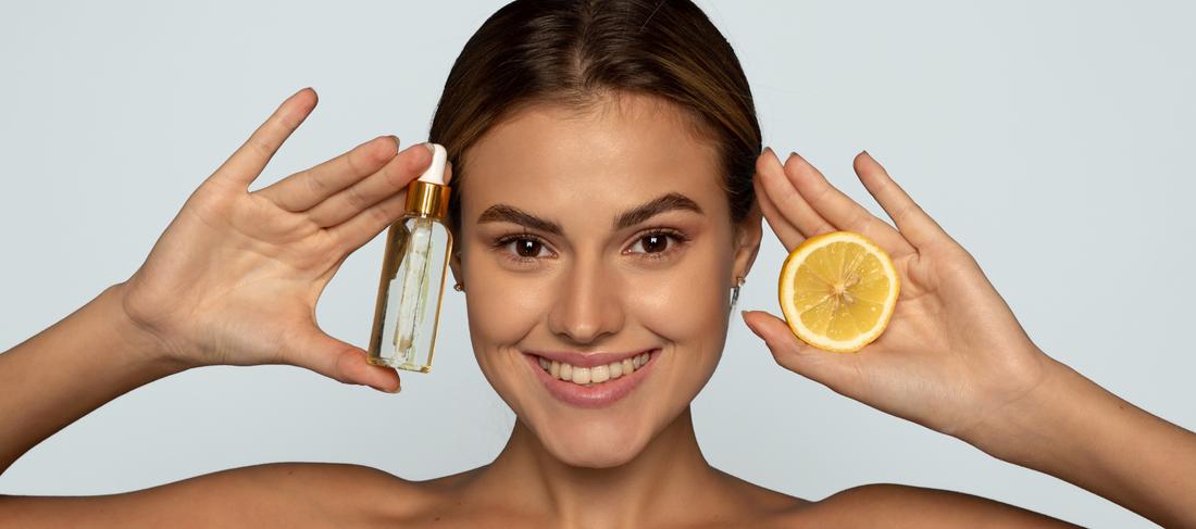 A young woman holds a dropper bottle of face serum in one hand and a lemon wedge in the other.