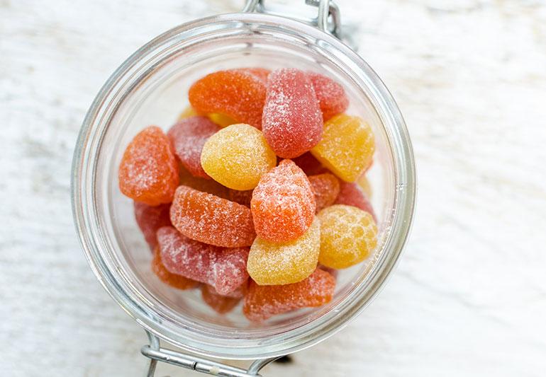 A jar of colorful fruit pectin gummies on a whitewashed wooden surface.