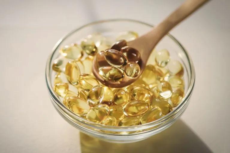 A wooden spoon holds several golden fish oil capsules above a glass bowl full of the same capsules.