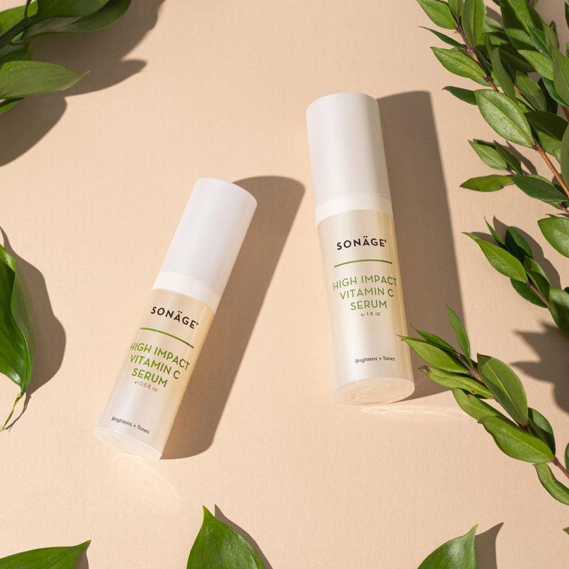 Two bottles of Sonäge High Impact Vitamin C Serum are placed on a beige surface, surrounded by green leaves.