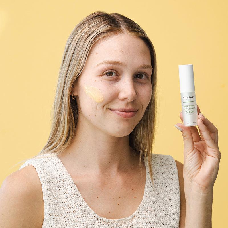 A young woman with blonde hair smiles at the camera while holding a bottle of vitamin C serum.