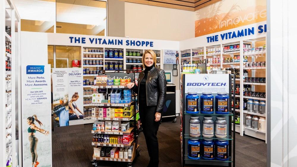 A woman in a black jacket stands in a store aisle with shelves of vitamins and supplements behind her.