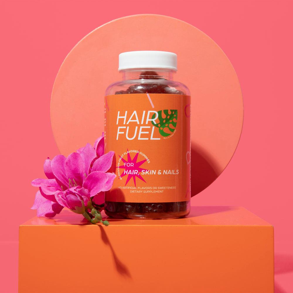 A pink and orange bottle of Hair Fuel gummies in front of a pink flower on an orange podium.