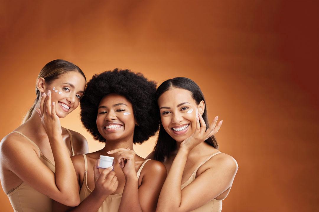 Three smiling women of different ethnicities are applying face cream to their cheeks.