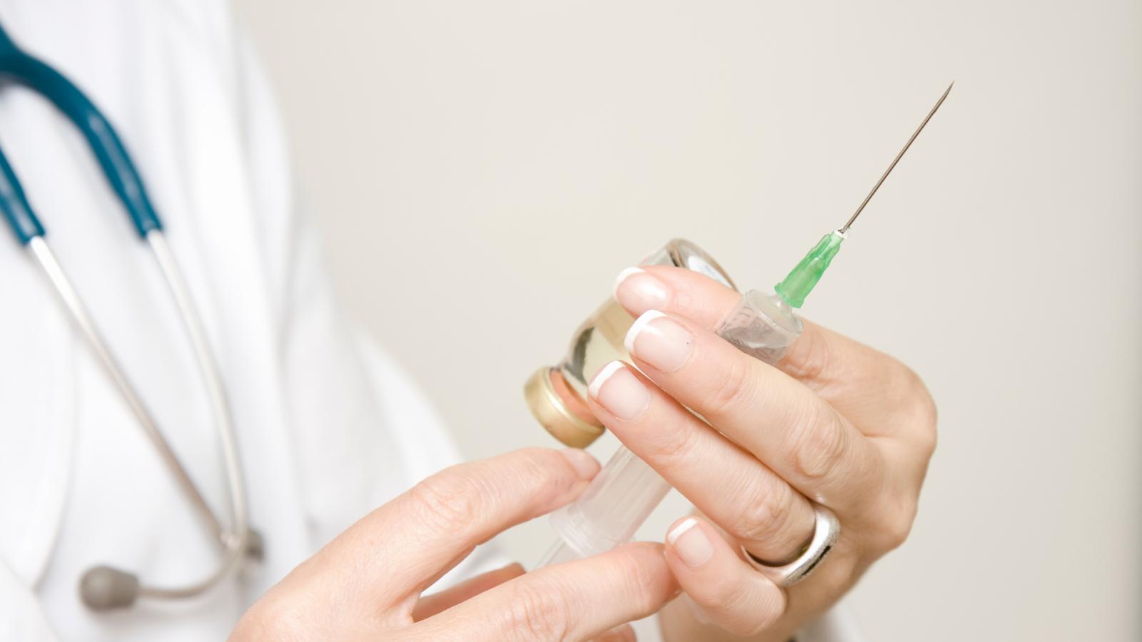A doctor in a white coat is holding a syringe with a needle and a vial with a green liquid.