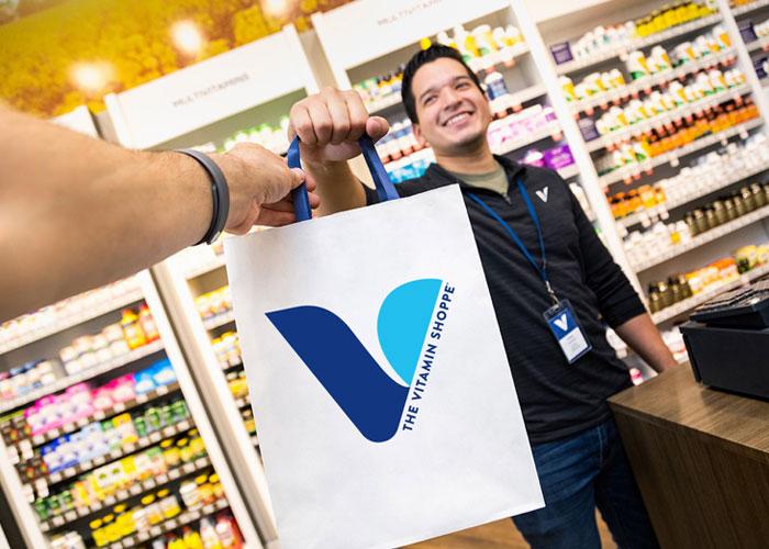 The Vitamin Shoppe employee is handing a white paper bag to a customer.