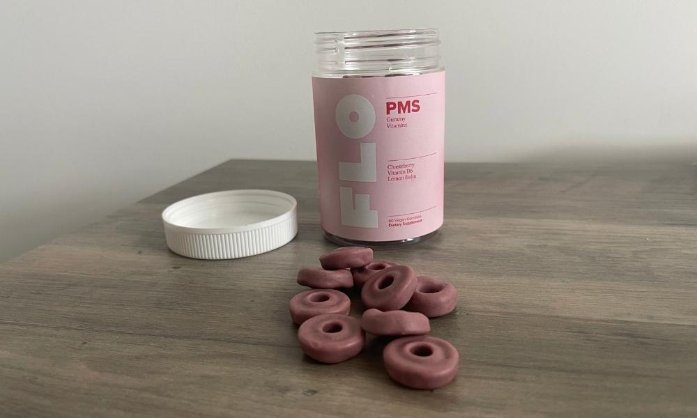 A clear jar of Flo PMS Vitamins with a white lid sits on a table next to several loose gummies in the shape of rings.