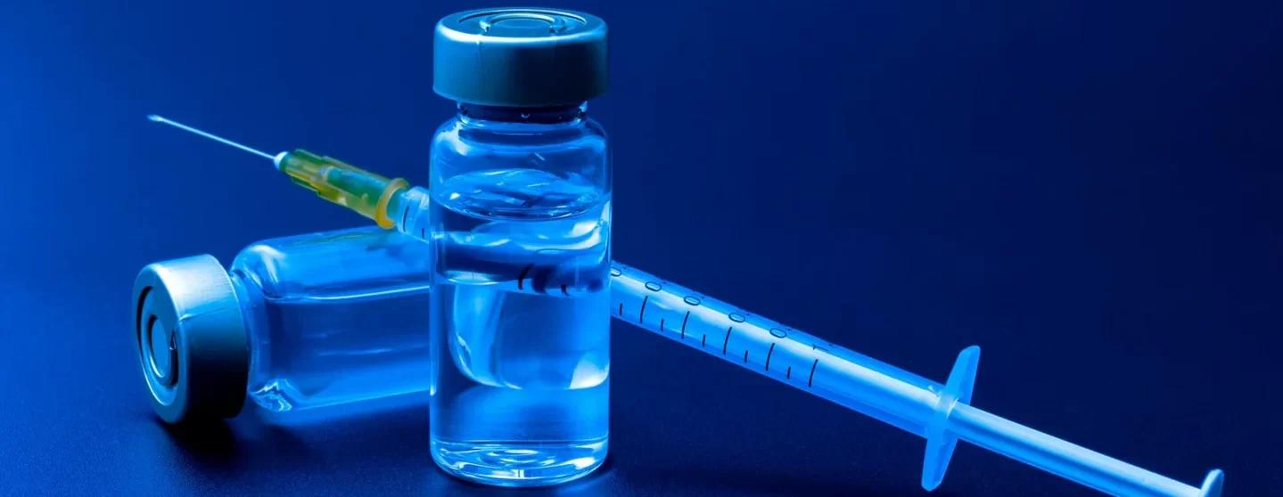 A close-up of a syringe and two vials with a blue liquid on a dark blue background.