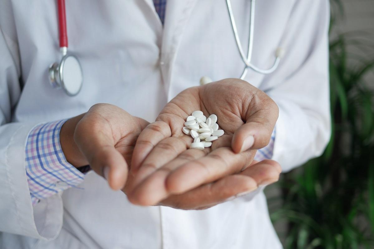 A doctor wearing a white coat and stethoscope holds a handful of white pills in their hand.
