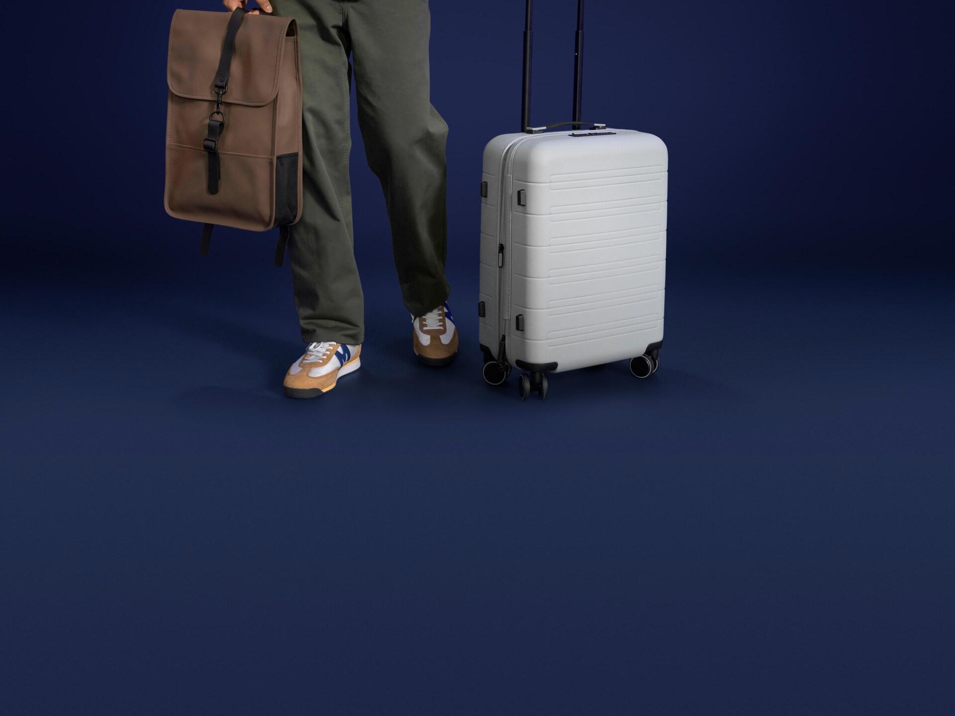 A person wearing sneakers, khaki pants, and a brown bag stands next to a silver suitcase.