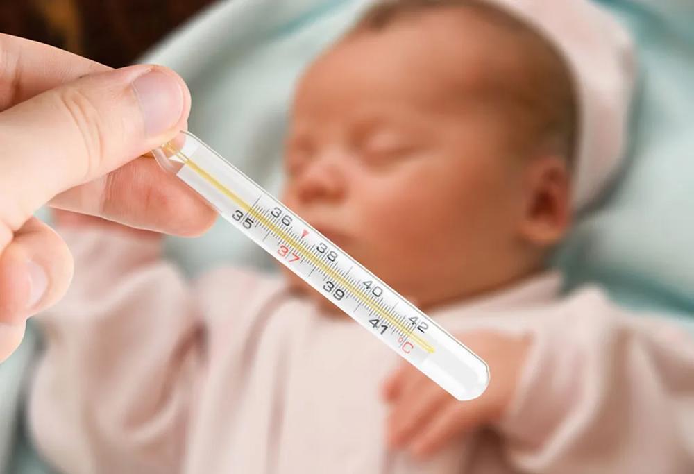 A hand holds a thermometer next to a sleeping babys head.