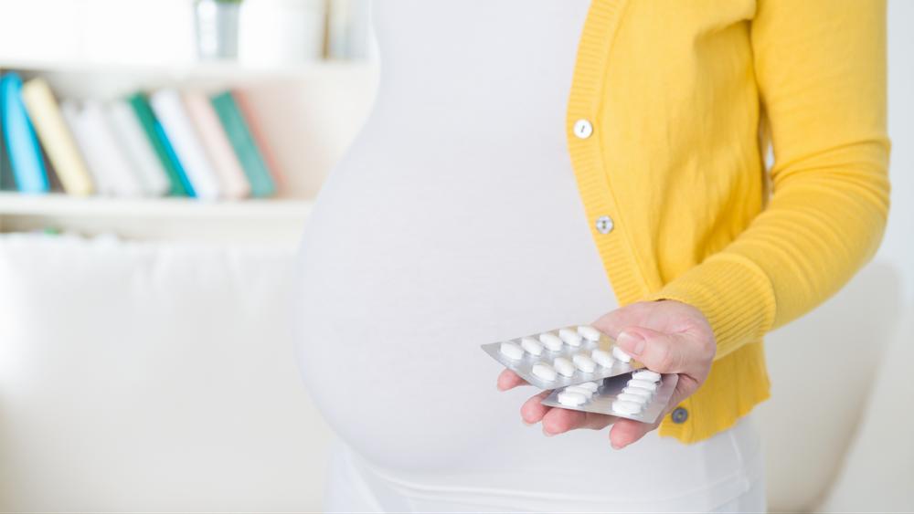 A pregnant woman wearing a yellow cardigan holds two blister packs of pills in her hand.