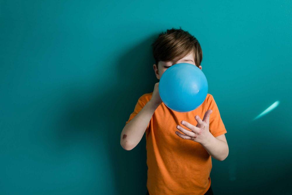 A boy in an orange shirt is blowing up a blue balloon.