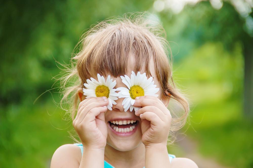 Little girl holding two daisies over her eyes.