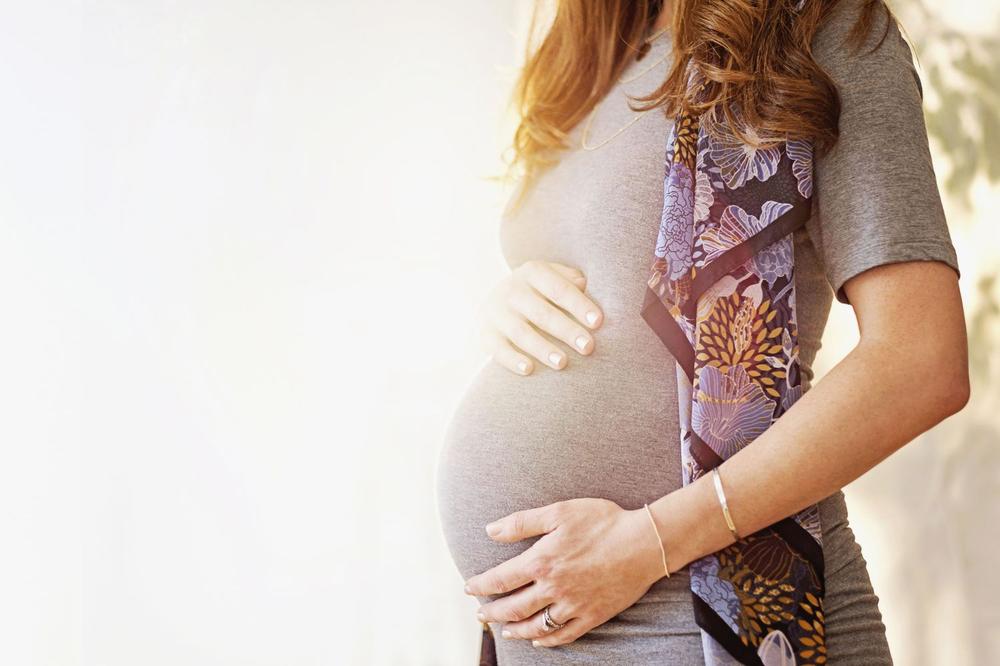 A pregnant woman wearing a gray dress with a floral scarf stands with her hand on her belly.