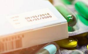 A close up of a white box with green pills spilled in front of it, with a label showing an expiration date.