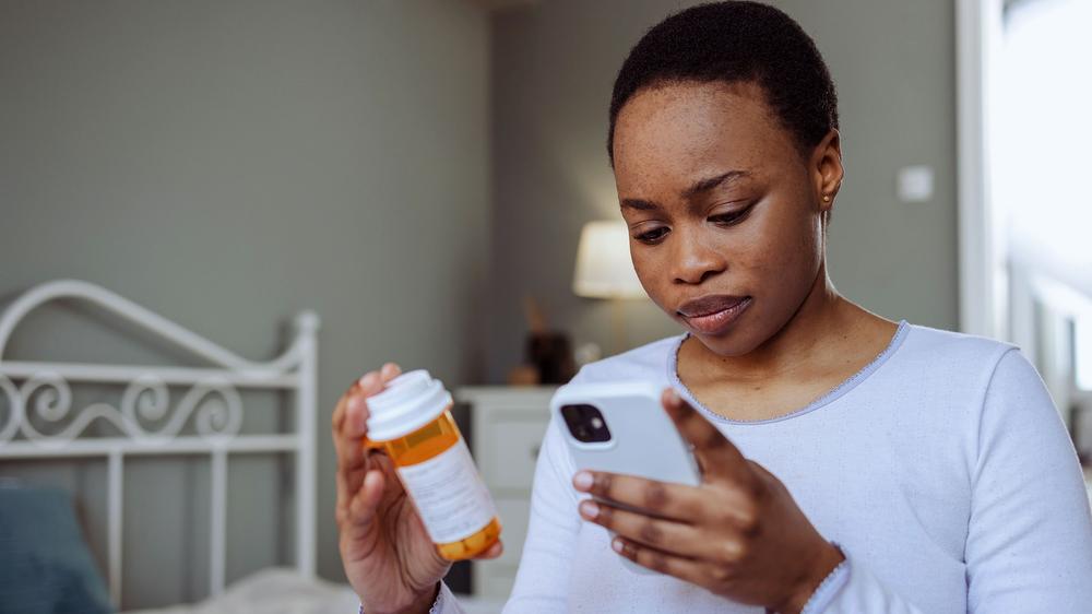 A young woman is holding a pill bottle and looking at her phone.