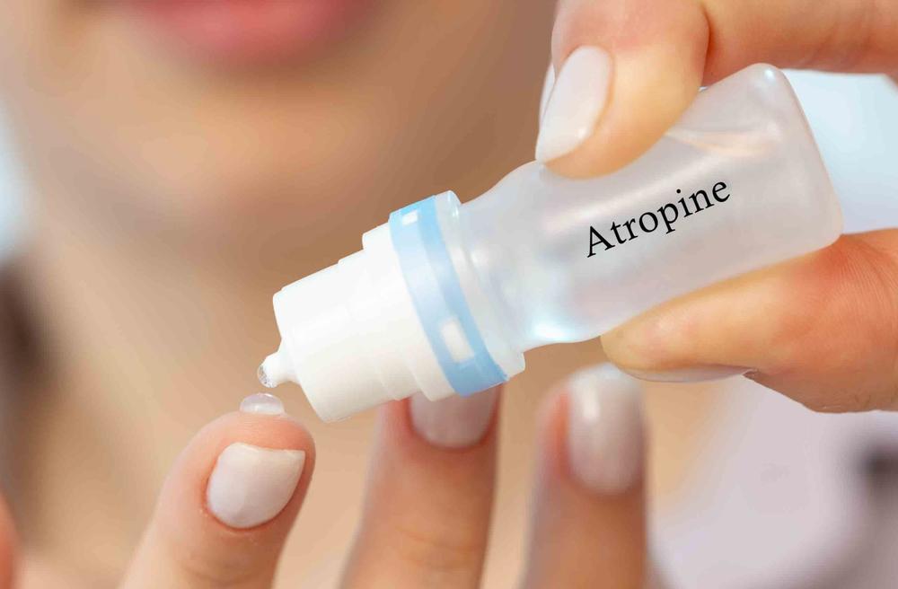 A woman is holding a bottle of eye drops labeled Atropine and putting a drop on her finger.