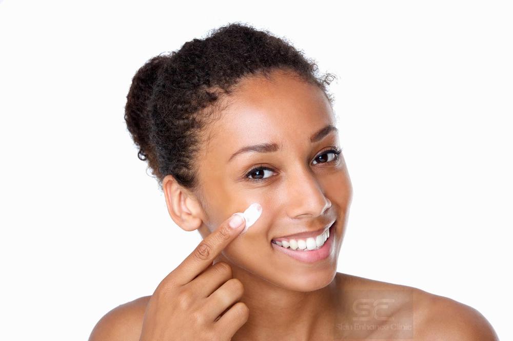 A young woman with dark skin is applying a white cream to her cheek and smiling.