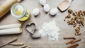 Various baking ingredients are arranged on a table, including flour, eggs, a rolling pin, cookie cutters, and cinnamon sticks.