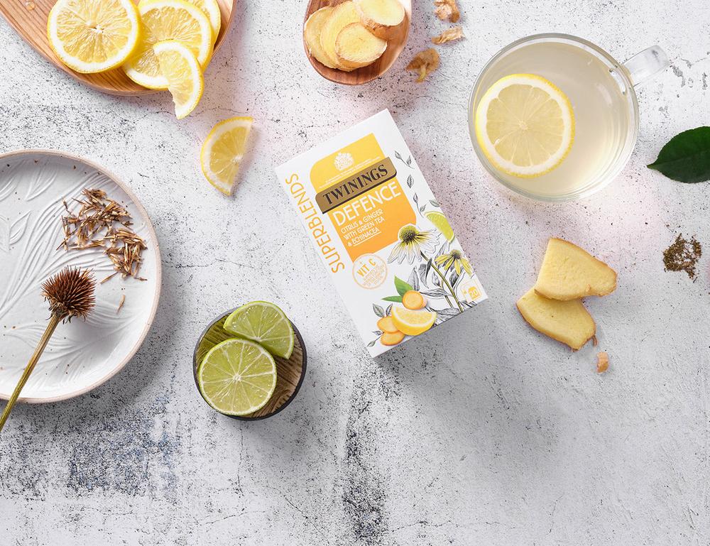 A box of Twinings Superblends Defence tea sits on a table scattered with lemon slices, lime wedges, and pieces of ginger.