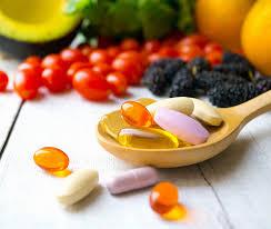 An assortment of colorful pills and capsules are on a wooden spoon in front of a pile of fruits and vegetables.