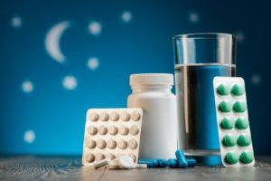 A bottle of pills, a glass of water, and two blister packs of pills on a table against a blue background with a crescent moon and stars.