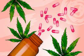 A brown bottle lies on its side with several pink pills spilling out onto green cannabis leaves.