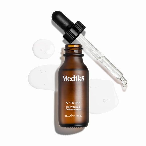 Close-up of a brown Medik8 bottle of C-Tetra Lipid Vitamin C Radiance Serum with a black dropper, and a clear liquid drop falling from the dropper.