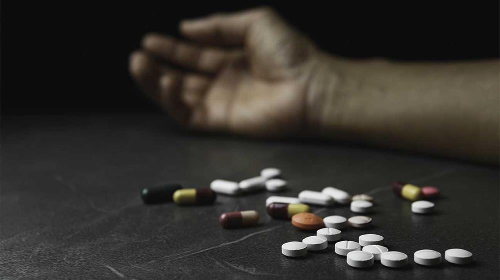 A hand lies limply on a table next to a pile of pills.