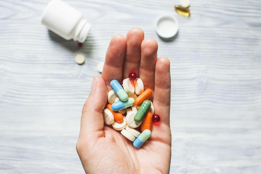 A handful of pills and capsules of various colors is held out in an open palm.