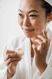 A young woman in a white bathrobe is smiling and applying cream to her face.