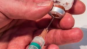 A gloved hand holding a syringe is shown preparing a dose of a vaccine.