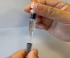 A gloved hand holding a syringe is shown injecting a fluid into a vial.