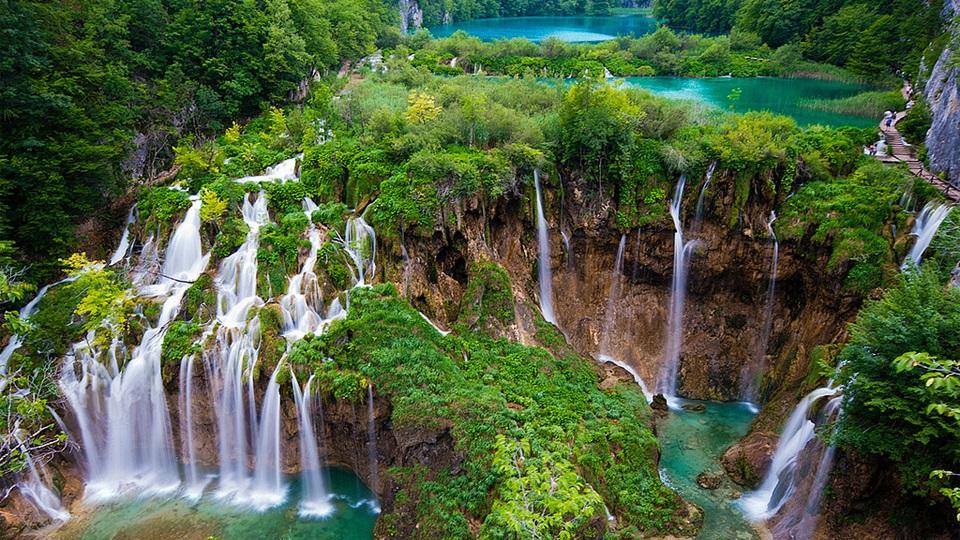 A photo of a lush green forest with waterfalls cascading over rocks into a turquoise lake.
