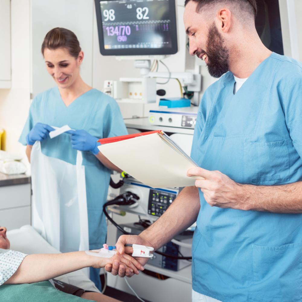 Bearded doctor in blue scrubs checks a patients pulse while a nurse in the background prepares an endoscope.