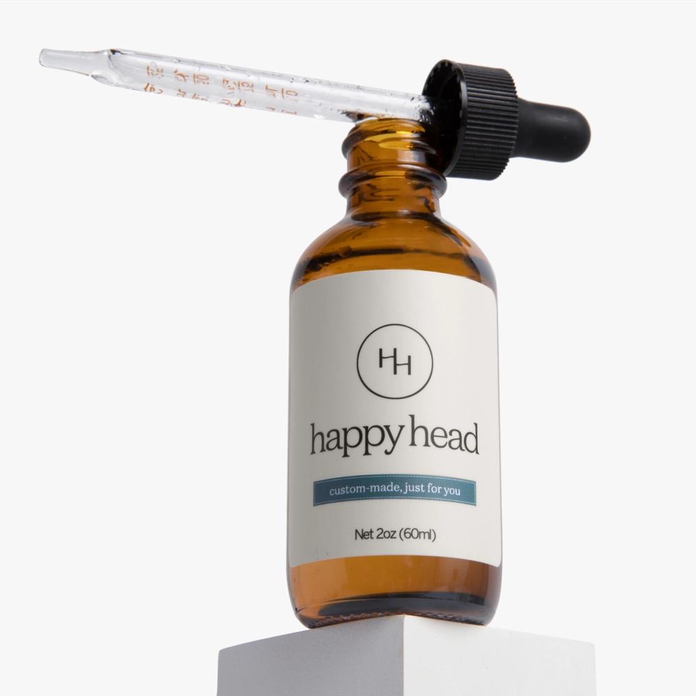 A brown bottle of Happy Head custom-made hair oil with a black dropper cap sits on a white pedestal.