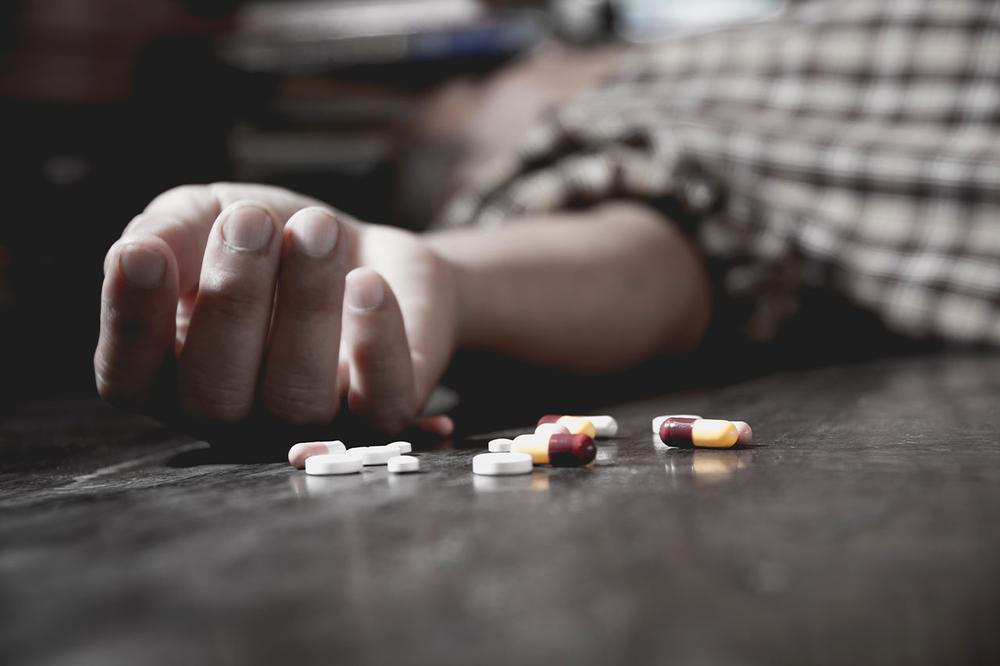 A hand lies motionless on a table next to scattered pills.